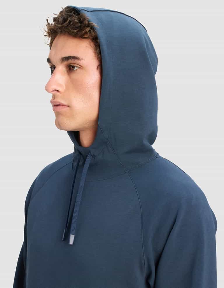 A young man with curly brown hair wears the Cozy Earth Men's StretchTech Hoodie with the hood up, looking to the left against a plain light gray background.|Color:Eclipse