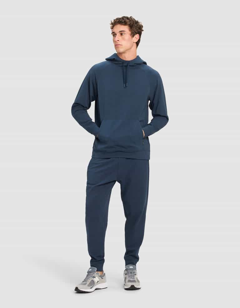 A man stands slightly to the side, wearing the Cozy Earth Men's StretchTech Hoodie & Jogger Set in dark blue with grey and white sneakers, posed against a plain light background.|Color:Eclipse