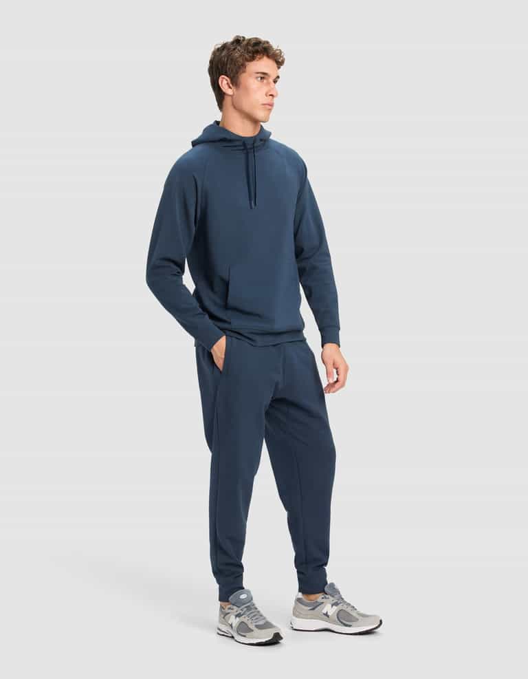 A man wears the Cozy Earth Men's StretchTech Hoodie & Jogger Set in navy, styled with gray and white sneakers, against a plain light gray background.|Color:Eclipse