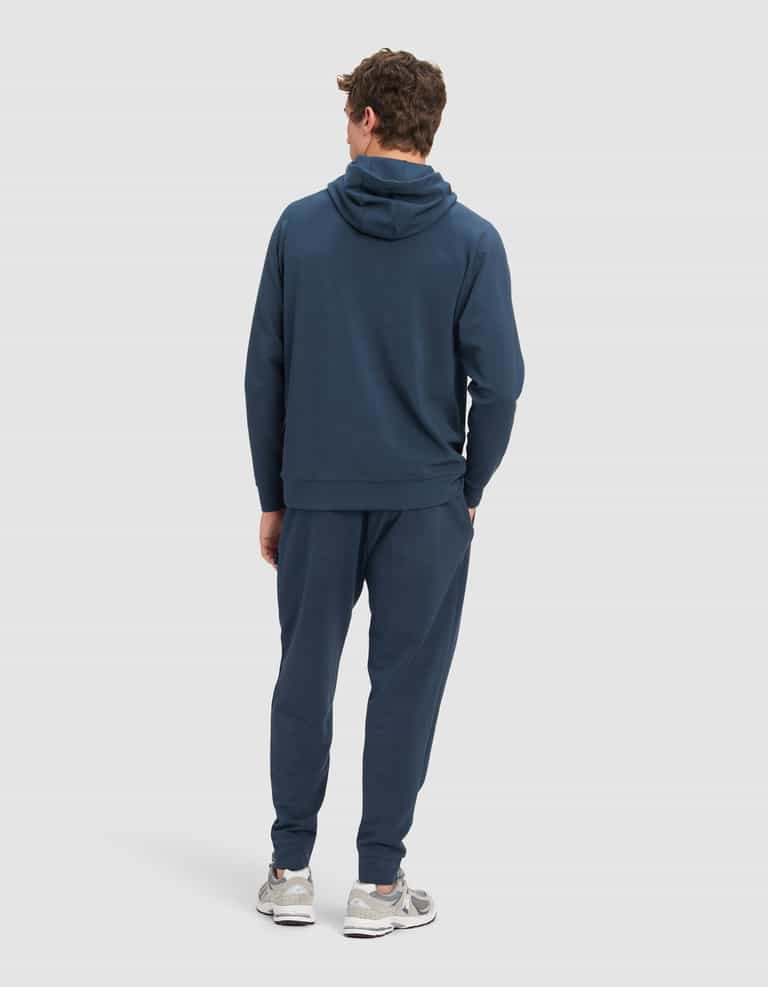 A man faces away, wearing the Cozy Earth Men's StretchTech Hoodie & Jogger Set in dark blue with light gray sneakers, against a plain light background.|Color:Eclipse