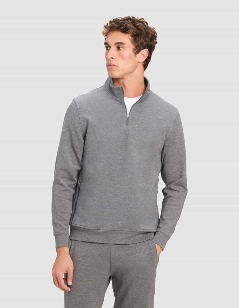 A young man wearing Cozy Earth's Men's StretchTech Quarter Zip and matching pants stands with one hand in his pocket, looking off to the side against a plain light gray background.|Color:Coal Heather
