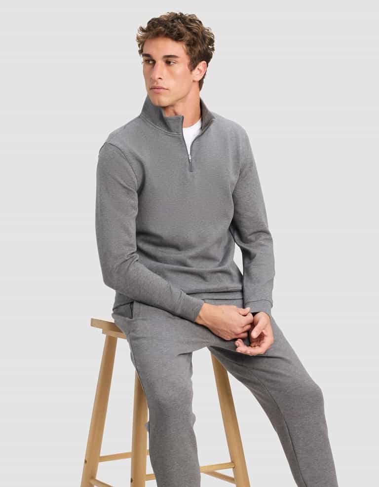 A young man wearing Cozy Earth's Men's StretchTech Quarter Zip in gray with matching pants sits on a wooden stool, looking to the side against a plain light gray background.|Color:Coal Heather