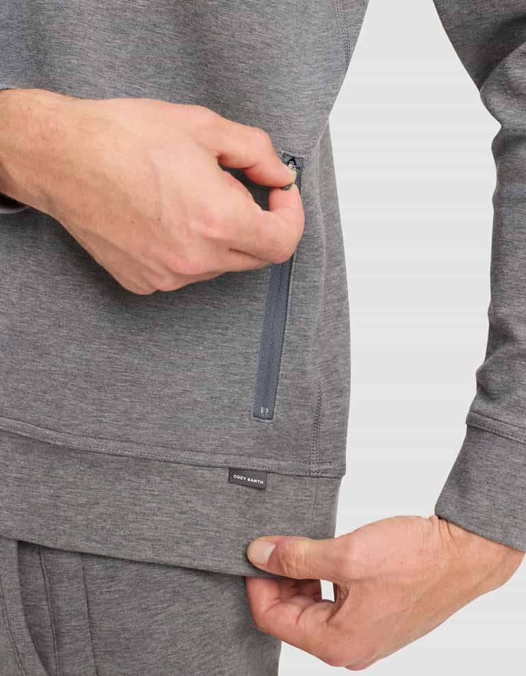 A person in a gray Men's StretchTech Quarter Zip by Cozy Earth is unzipping a vertical side pocket while holding the hem with the other hand. The sweatshirt features a small black "Cozy Earth" label.|Color:Coal Heather