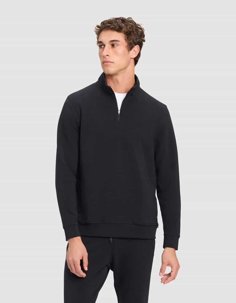 A young man in Cozy Earth's Men's StretchTech Quarter Zip and matching black pants stands against a light gray background, looking slightly to the side with a neutral expression.|Color:Jet Black
