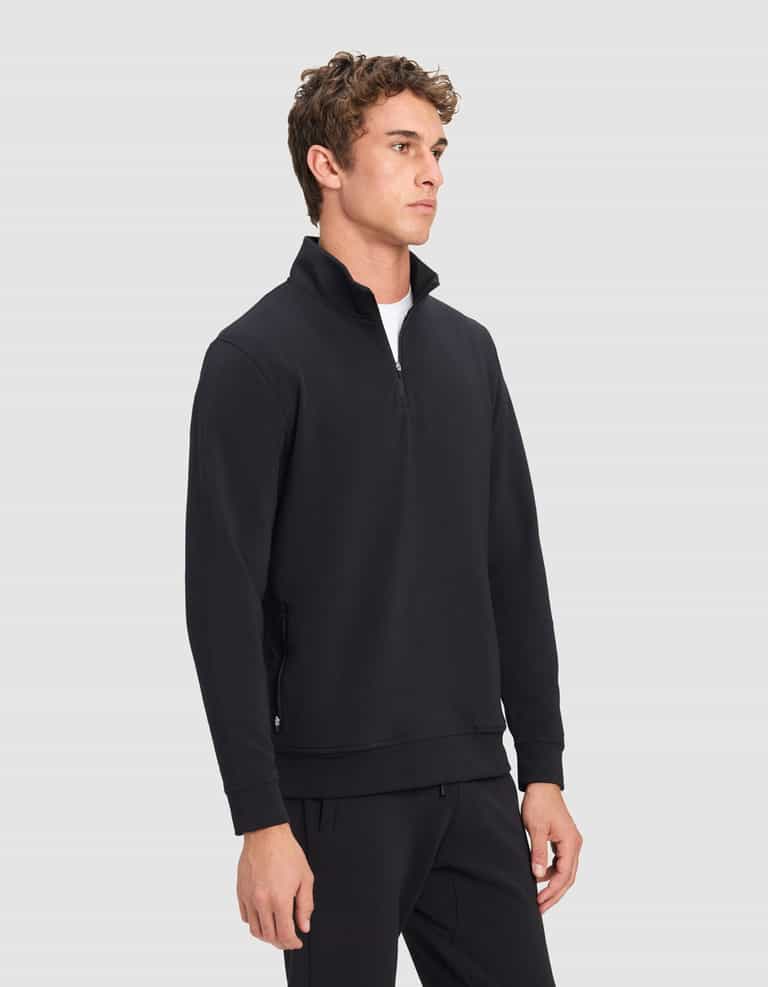 A young man stands slightly turned against a plain light gray background, wearing a Cozy Earth Men's StretchTech Quarter Zip pullover with matching black pants.|Color:Jet Black