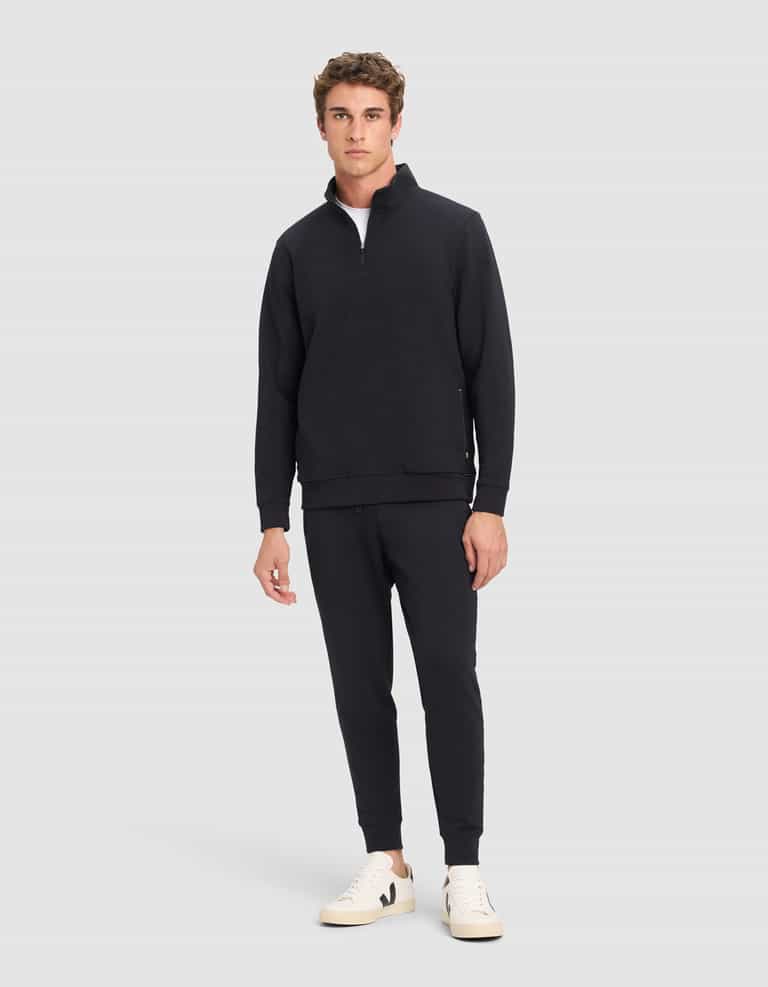 A man stands against a plain background wearing Cozy Earth's Men's StretchTech Quarter Zip in black, matching black joggers, and white sneakers. He faces forward with a neutral expression and relaxed posture.|Color:Jet Black