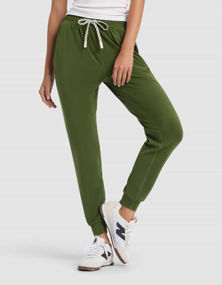 A woman stands against a plain light background wearing a white top, Cozy Earth Women's Studio Jogger pants in olive green with a drawstring, and white New Balance sneakers, one leg slightly bent.|Color:Grove