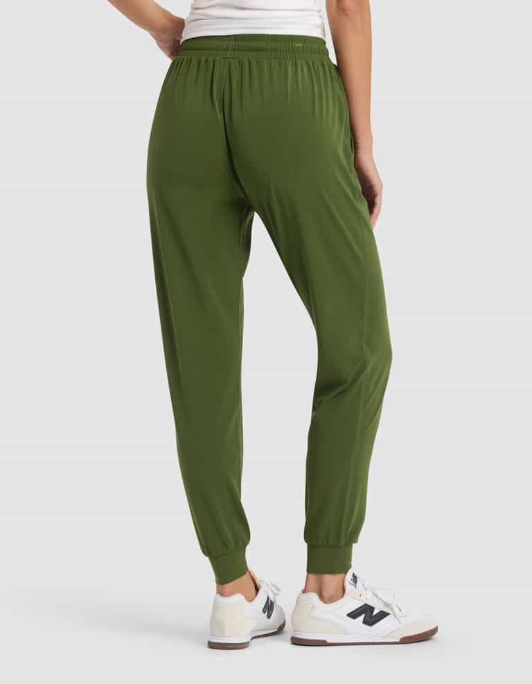 A woman is shown from the waist down wearing Cozy Earth Women's Studio Jogger pants in olive green, paired with white sneakers, standing on a light gray background.|Color:Grove