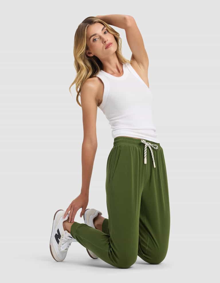 A woman with long, wavy blonde hair kneels on a light gray background, wearing Cozy Earth's Women's Studio Jogger in green, a white sleeveless top, and white sneakers. She poses with one hand behind her head.|Color:Grove