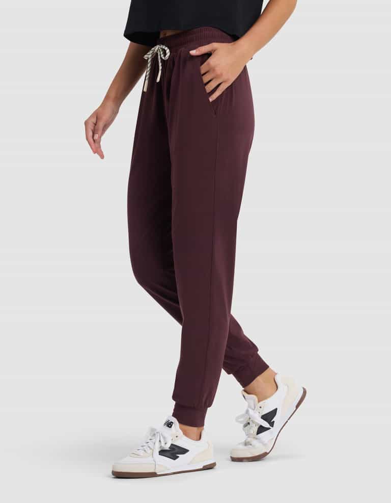 A person models the Cozy Earth Women's Studio Jogger in burgundy, paired with a black top and white sneakers with black accents, standing with one hand in a pocket against a plain light background.|Color:Raisin