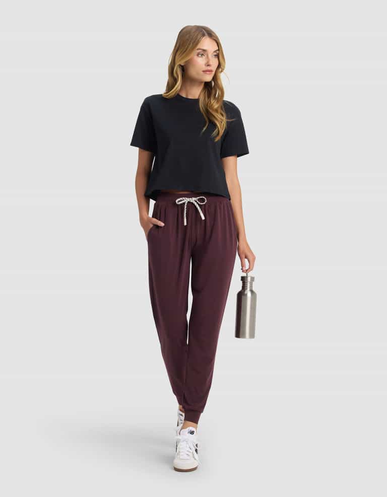 A woman with long hair models Cozy Earth's Women's Studio Jogger in maroon, paired with a black t-shirt and white sneakers. She holds a stainless steel water bottle and looks to the side against a plain light gray background.|Color:Raisin