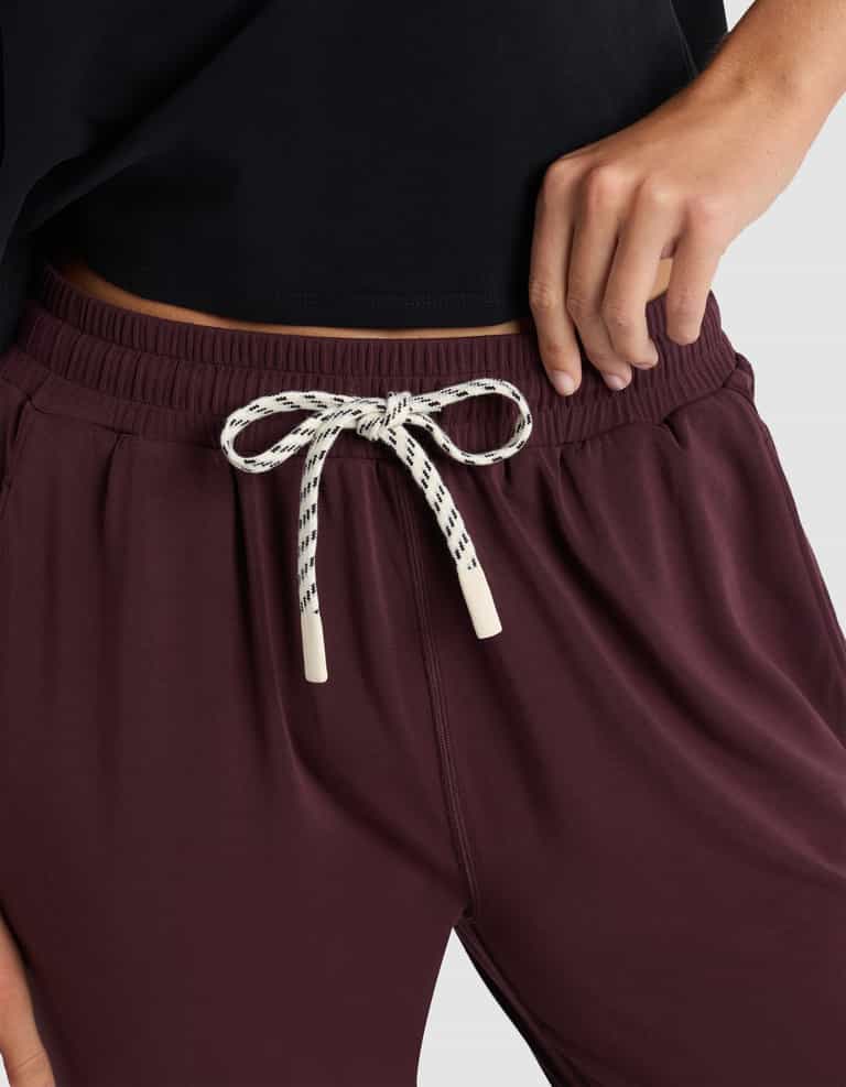 A woman models the Cozy Earth Women's Studio Jogger in maroon, featuring a white and black patterned drawstring tied in a bow, paired with a black top and posing with one hand on her hip.|Color:Raisin