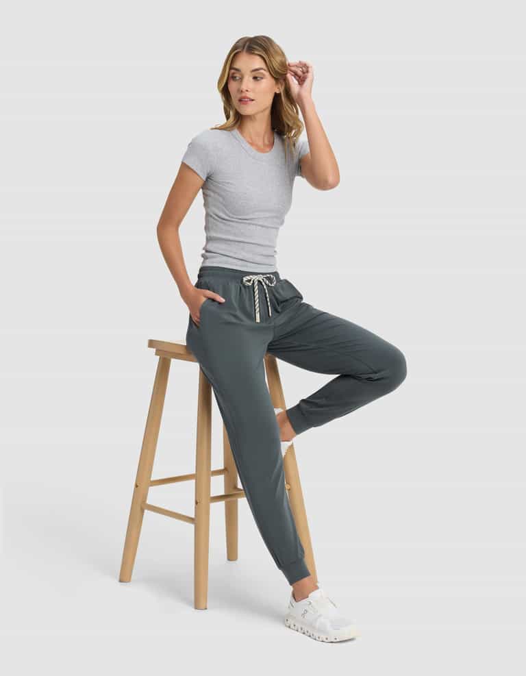 A woman in Cozy Earth Women's Studio Jogger and a fitted gray t-shirt sits on a wooden stool against a light background, wearing white sneakers, one hand in her pocket and the other touching her hair.|Color:Storm
