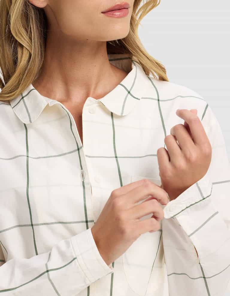 A woman with blonde hair adjusts the cuff of her white, green-plaid Women's Sutton Long Sleeve Pajama Set by Cozy Earth. Her face is partly visible against a plain light gray background.|Color:Bone Windowpane