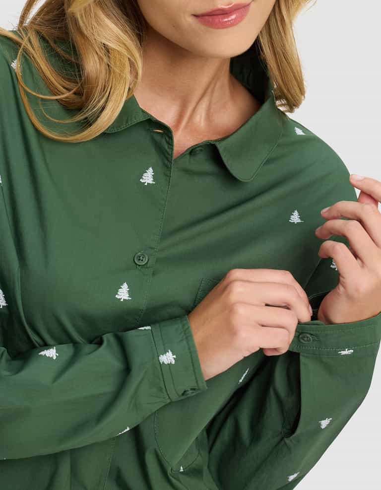 A woman wearing the Cozy Earth Women's Sutton Long Sleeve Pajama Set in green, featuring small white tree patterns, gently touches her sleeve. Only the lower half of her face and her loose light brown hair are visible.|Color:Jade Tree