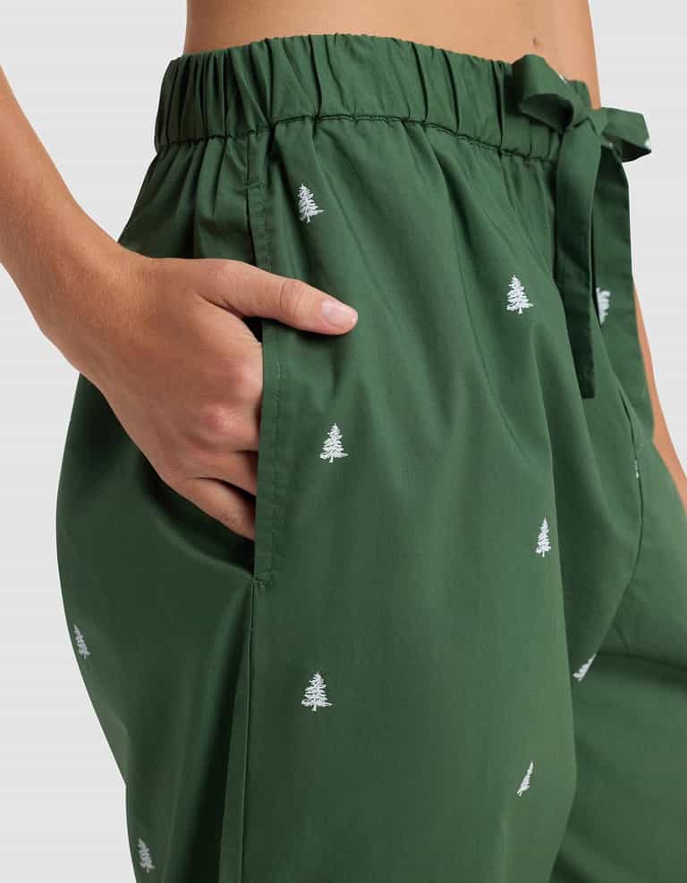 Close-up of a person wearing Cozy Earth's Women's Sutton Long Sleeve Pajama Set featuring green pants with white embroidered trees, an elastic waistband, and a fabric tie belt, their right hand resting in the pocket.|Color:Jade Tree
