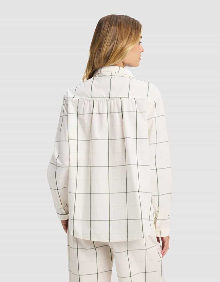 A woman with light brown hair is seen from the back wearing the Cozy Earth Women's Sutton Long Sleeve Pajama Set—a loose-fitting white top and matching pants with a thin black grid pattern—against a plain, light background.|Color:Bone Windowpane