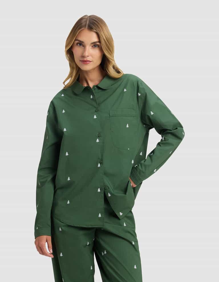 A woman with wavy blonde hair models the Cozy Earth Women's Sutton Long Sleeve Pajama Set—dark green with a white tree print—standing with one hand in her pocket against a plain light background.|Color:Jade Tree