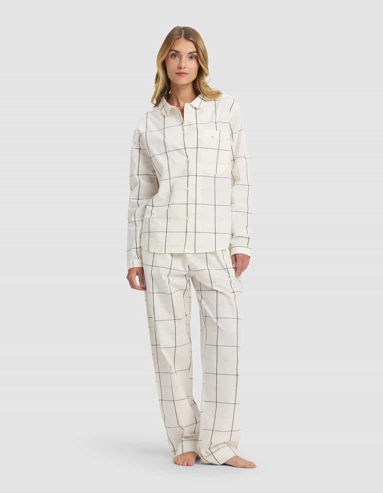 A woman models the Cozy Earth Women's Sutton Long Sleeve Pajama Set, featuring a white button-up top and loose pants with a black grid pattern, standing barefoot against a plain light gray background.|Color:Bone Windowpane