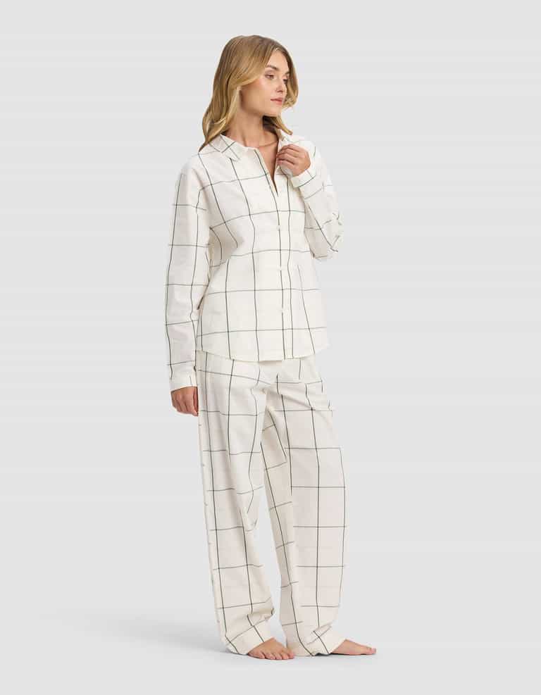 A woman poses barefoot against a plain light background wearing the Cozy Earth Women's Sutton Long Sleeve Pajama Set, featuring a matching white button-up shirt and loose-fitting pants with a subtle black grid pattern.|Color:Bone Windowpane