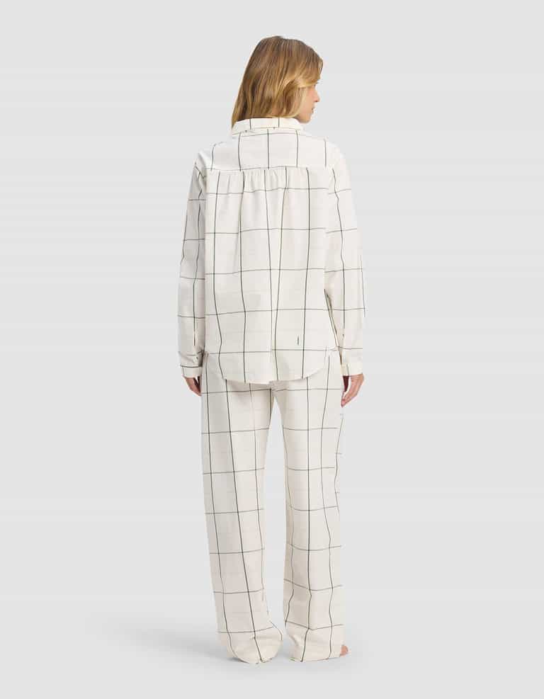 A person faces away, wearing the Cozy Earth Women's Sutton Long Sleeve Pajama Set—white with thin black plaid lines forming large squares. The set includes a loose-fitting top and pants. Background is plain light gray.|Color:Bone Windowpane