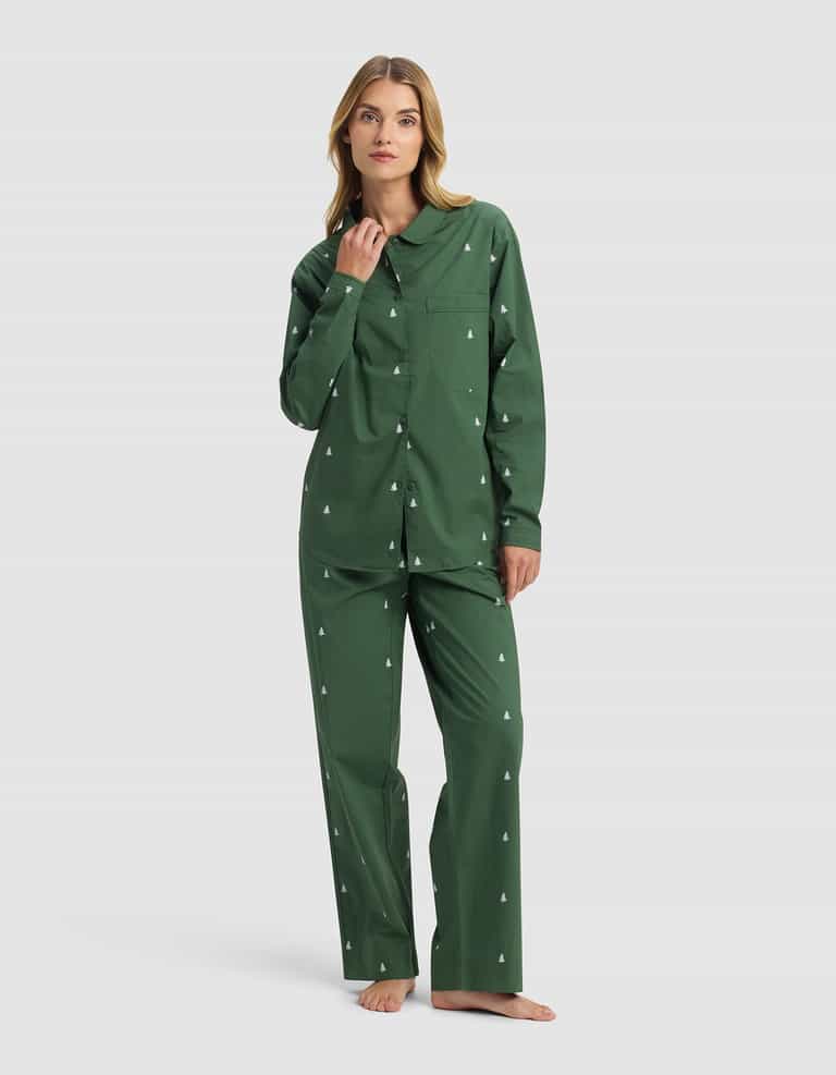 A woman stands barefoot in Cozy Earth's Women's Sutton Long Sleeve Pajama Set, featuring green pajamas with a small white tree pattern, a button-up long-sleeve shirt, and matching pants, against a plain white background.|Color:Jade Tree