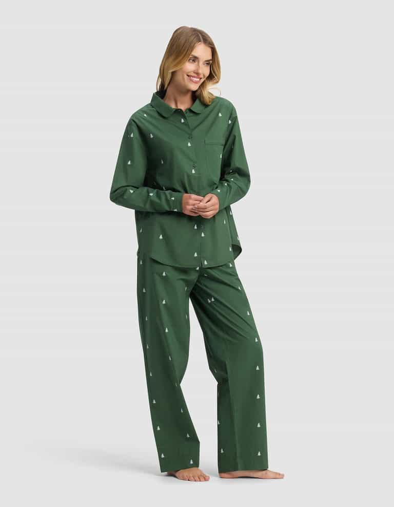 A woman stands barefoot and smiling against a plain light background, wearing Cozy Earth's Women's Sutton Long Sleeve Pajama Set in dark green with small white tree patterns.|Color:Jade Tree
