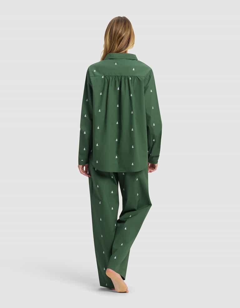 A person with long hair stands barefoot, facing away, wearing the Women's Sutton Long Sleeve Pajama Set by Cozy Earth in dark green with a small white tree pattern, set against a plain light background.|Color:Jade Tree