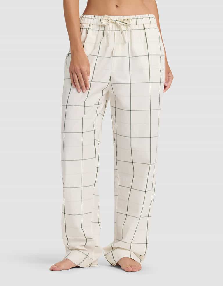 A woman stands barefoot in Cozy Earth’s Women's Sutton Long Sleeve Pajama Set—loose-fitting white pants with a green plaid pattern and drawstring waistband—against a plain, light gray background.|Color:Bone Windowpane