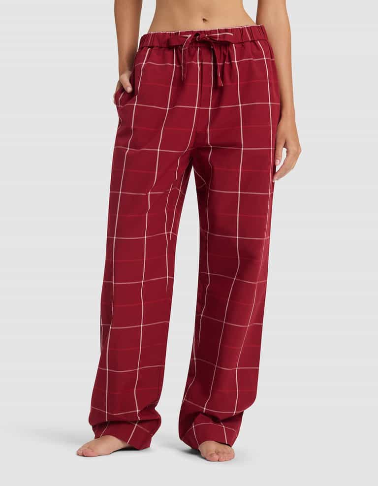 A woman wears Cozy Earth's Women's Sutton Long Sleeve Pajama Set featuring loose-fitting, red pajama pants with a white grid pattern, drawstring waist, and pockets, standing barefoot against a plain light background.|Color:Garnet Windowpane