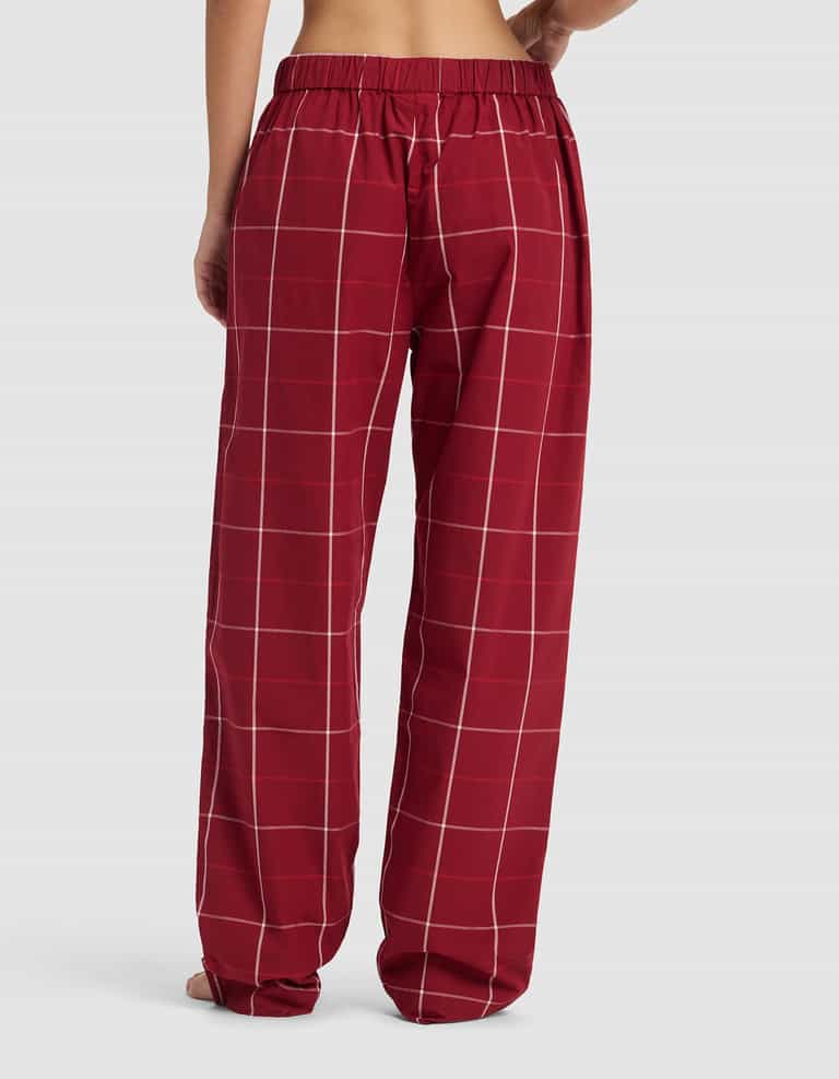 A woman is shown from the back wearing Cozy Earth's Women's Sutton Long Sleeve Pajama Set—loose-fitting red plaid pants with a white checkered pattern and an elastic waistband, barefoot against a plain light background.|Color:Garnet Windowpane