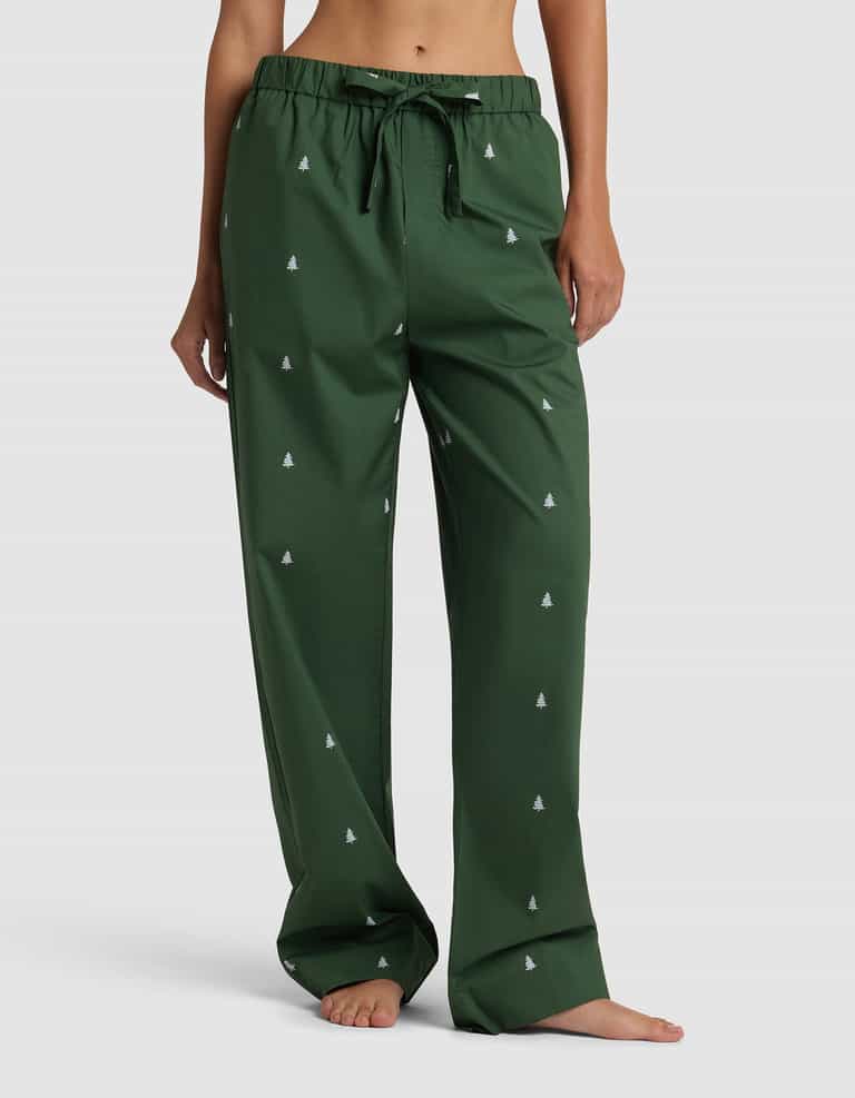 A person stands barefoot wearing the dark green pajama pants from Cozy Earth's Women's Sutton Long Sleeve Pajama Set, featuring a drawstring waist and small white tree patterns. The focus is on the pants; the upper body is mostly out of frame.|Color:Jade Tree