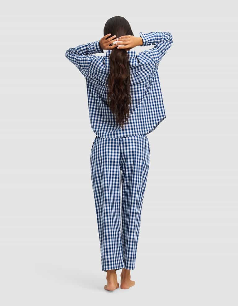 A woman with long, wavy hair stands barefoot facing away, stretching with her hands behind her head while wearing the Cozy Earth Women's Sutton Long Sleeve Pajama Set in blue and white checkered pattern against a light gray background.|Color:Pacific Gingham