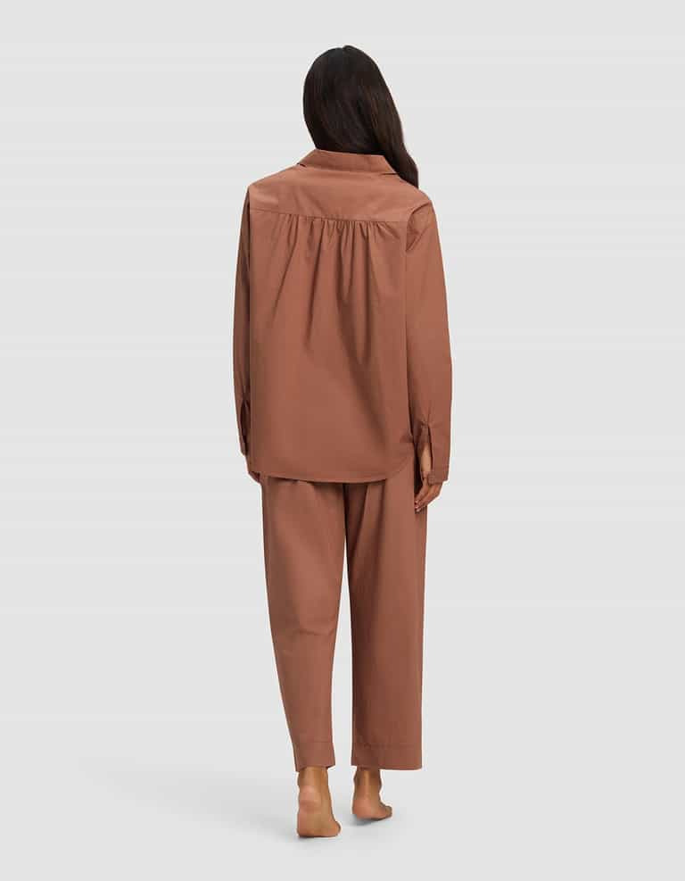 A barefoot woman with long dark hair is shown from behind wearing the Cozy Earth Women's Sutton Long Sleeve Pajama Set in brown, featuring a loose fit and long sleeves and pants, against a plain light gray background.|Color:Saddle