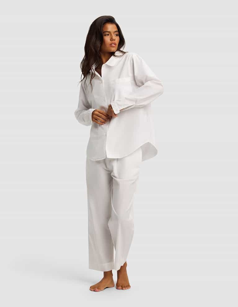 A woman with long dark hair stands barefoot against a light gray background, wearing the Cozy Earth Women's Sutton Long Sleeve Pajama Set—loose-fitting white pajamas featuring a button-up shirt and wide-leg pants.|Color:White