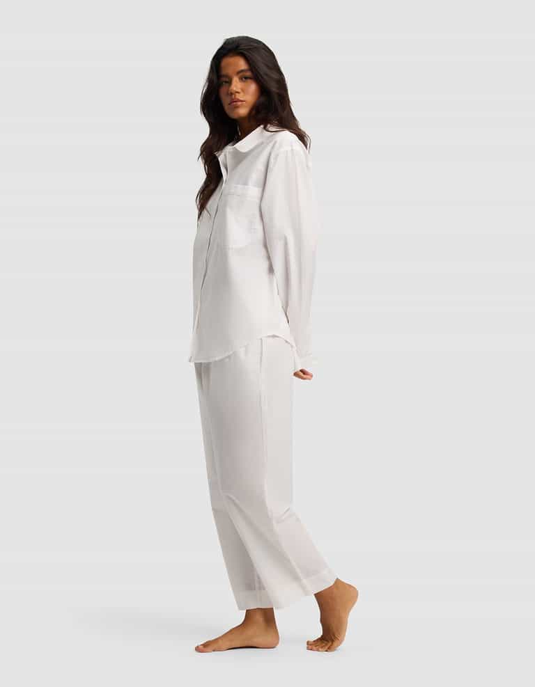 A woman with long dark hair stands barefoot in the Women's Sutton Long Sleeve Pajama Set by Cozy Earth—loose-fitting white shirt and matching pants—against a plain light gray background.|Color:White