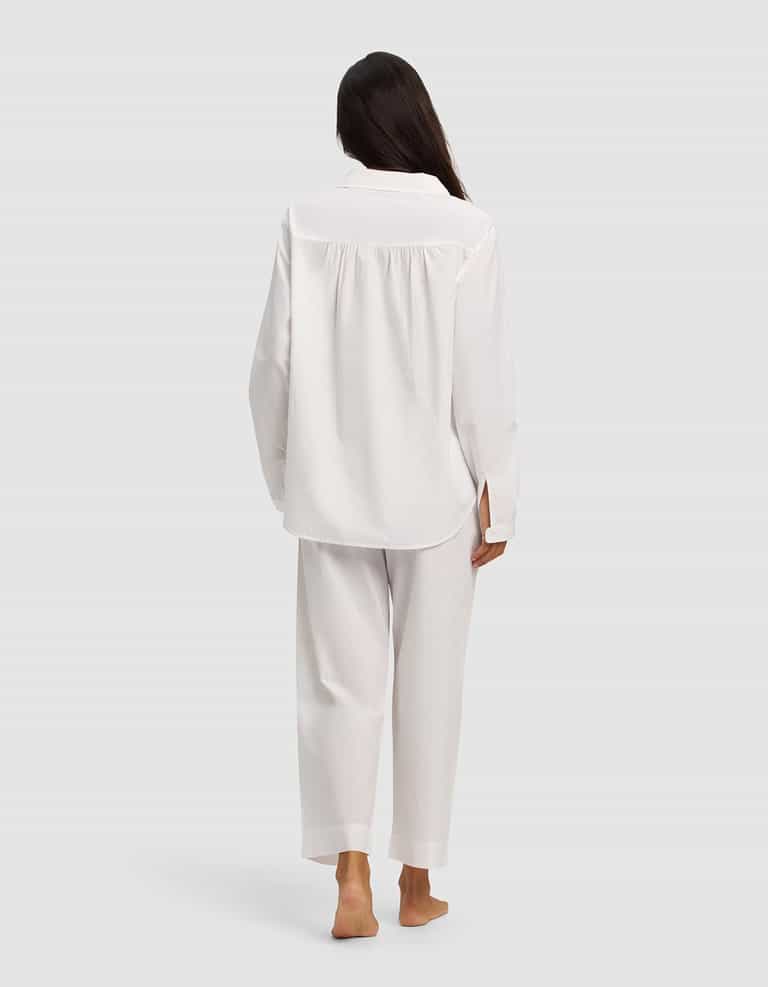 A woman with long dark hair, seen from behind, stands barefoot on a light gray background wearing the Cozy Earth Women's Sutton Long Sleeve Pajama Set in white.|Color:White
