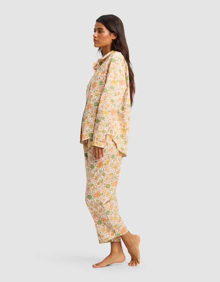 A woman stands sideways barefoot against a plain light gray background, wearing the Cozy Earth Women's Sutton Long Sleeve Pajama Set—loose-fitting, floral-patterned pajamas in shades of peach, yellow, and green.|Color:Wildflower