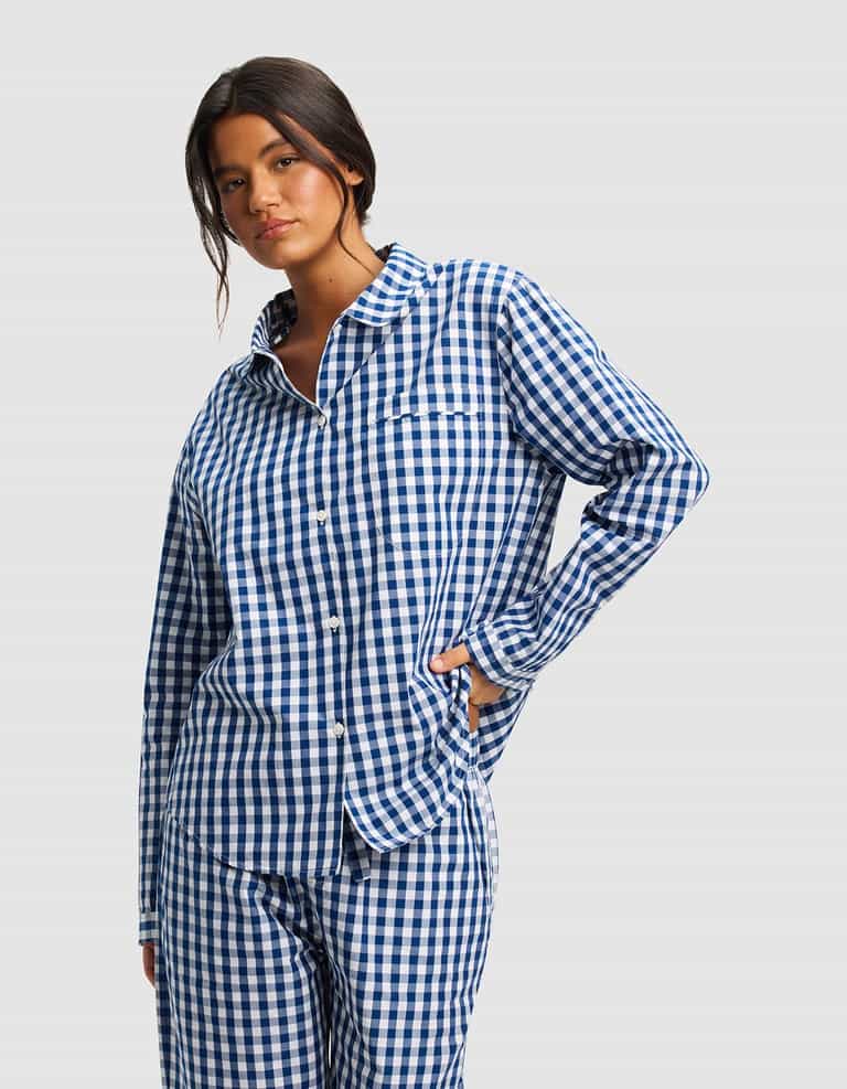 A woman models the Cozy Earth Women's Sutton Long Sleeve Pajama Top, featuring blue and white checks and matching pants, against a plain light gray background.|Color:Pacific Gingham