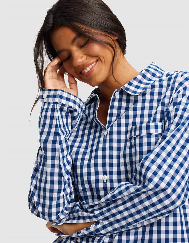 A woman with long dark hair smiles with her eyes closed, touching her head while wearing the Cozy Earth Women's Sutton Long Sleeve Pajama Top against a light gray background.|Color:Pacific Gingham