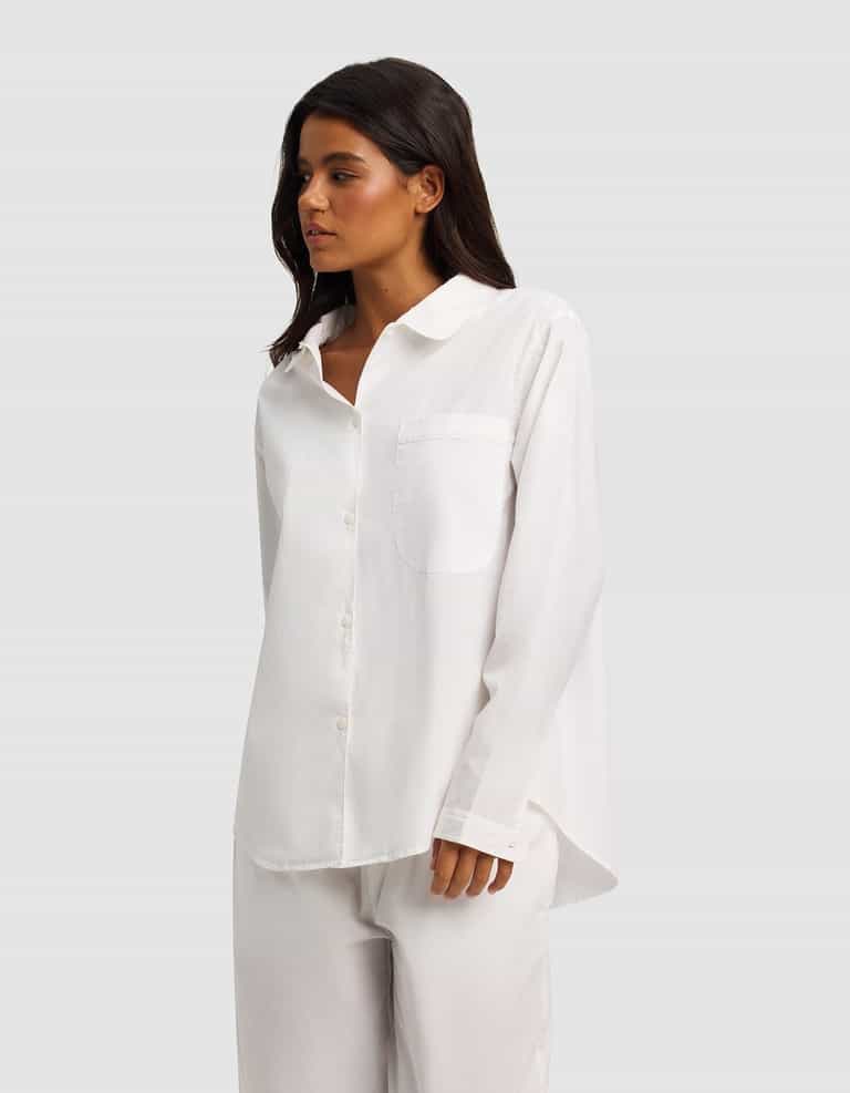 A woman with long dark hair wears the Cozy Earth Women's Sutton Long Sleeve Pajama Top— a loose-fitting white button-up with a chest pocket—paired with matching white pants, standing against a plain light gray background.|Color:White