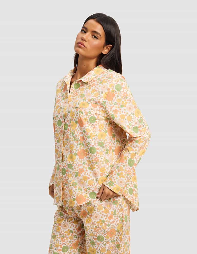 A woman with long dark hair models the Cozy Earth Women's Sutton Long Sleeve Pajama Top featuring a colorful floral print in orange, yellow, and green, standing with her hands in her pockets against a light gray backdrop.|Color:Wildflower