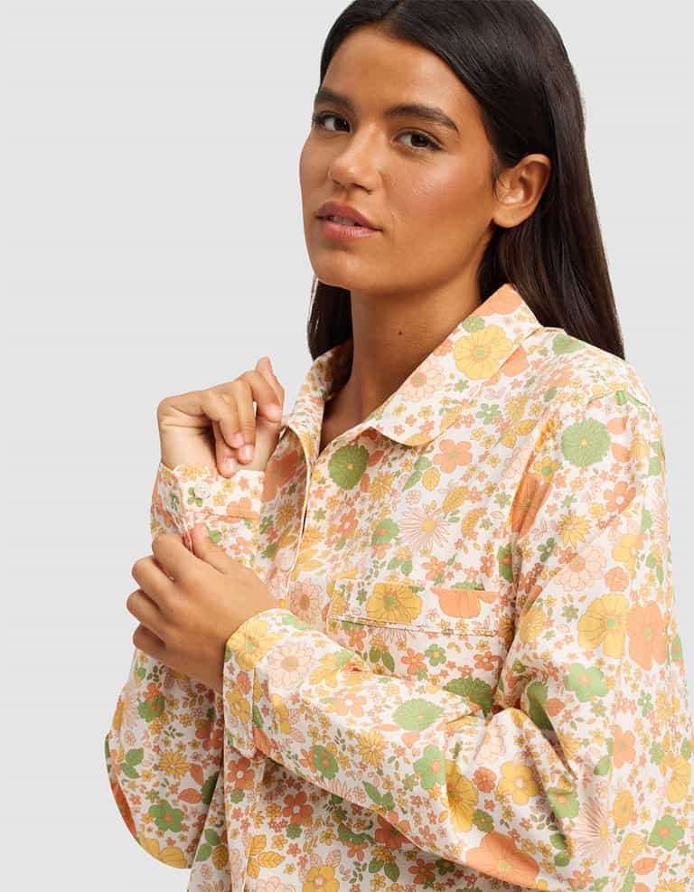 A woman with long dark hair wears the Women's Sutton Long Sleeve Pajama Top by Cozy Earth, featuring a colorful floral pattern in orange, yellow, and green, standing against a plain light gray background.|Color:Wildflower
