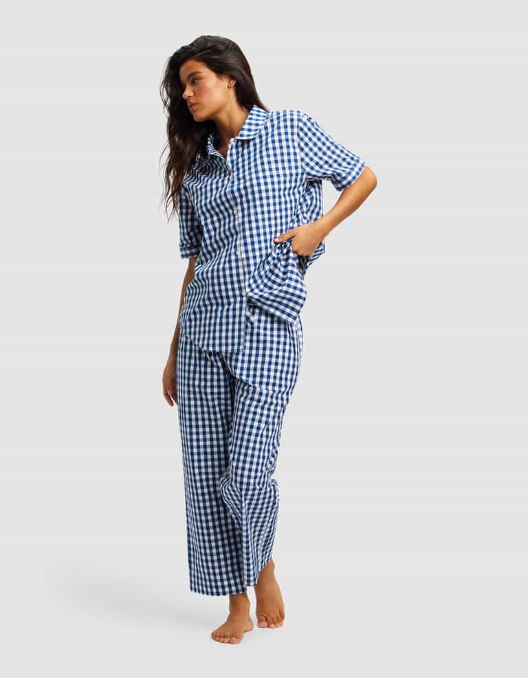 A woman stands barefoot against a plain light gray background, wearing Cozy Earth's Women's Sutton Cropped Pajama Pant—a blue and white checkered set with short sleeves and cropped pants.|Color:Pacific Gingham
