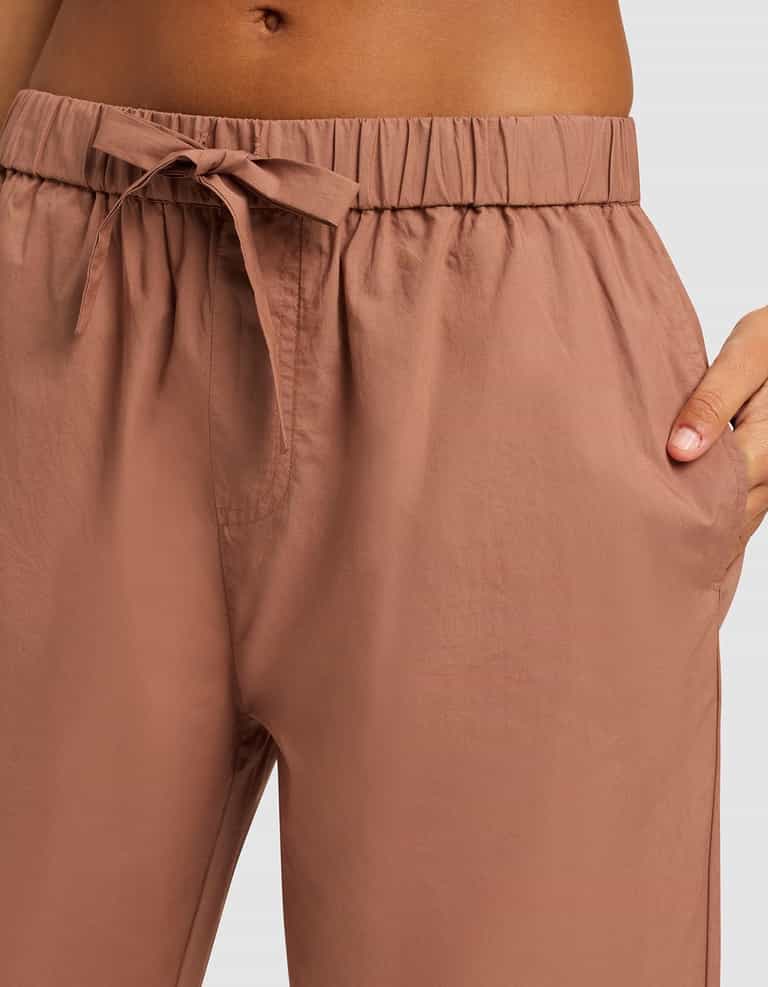 A close-up shows a person wearing Cozy Earth's Women's Sutton Cropped Pajama Pant in light brown, featuring a high waist, elastic waistband, drawstring tie, and their hand in one pocket.|Color:Saddle