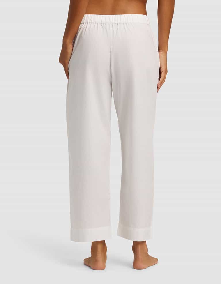 A woman is shown from the back wearing Cozy Earth's Women's Sutton Cropped Pajama Pant, featuring a loose fit, white color, elastic waistband, and side pockets; she is barefoot against a plain background.|Color:White