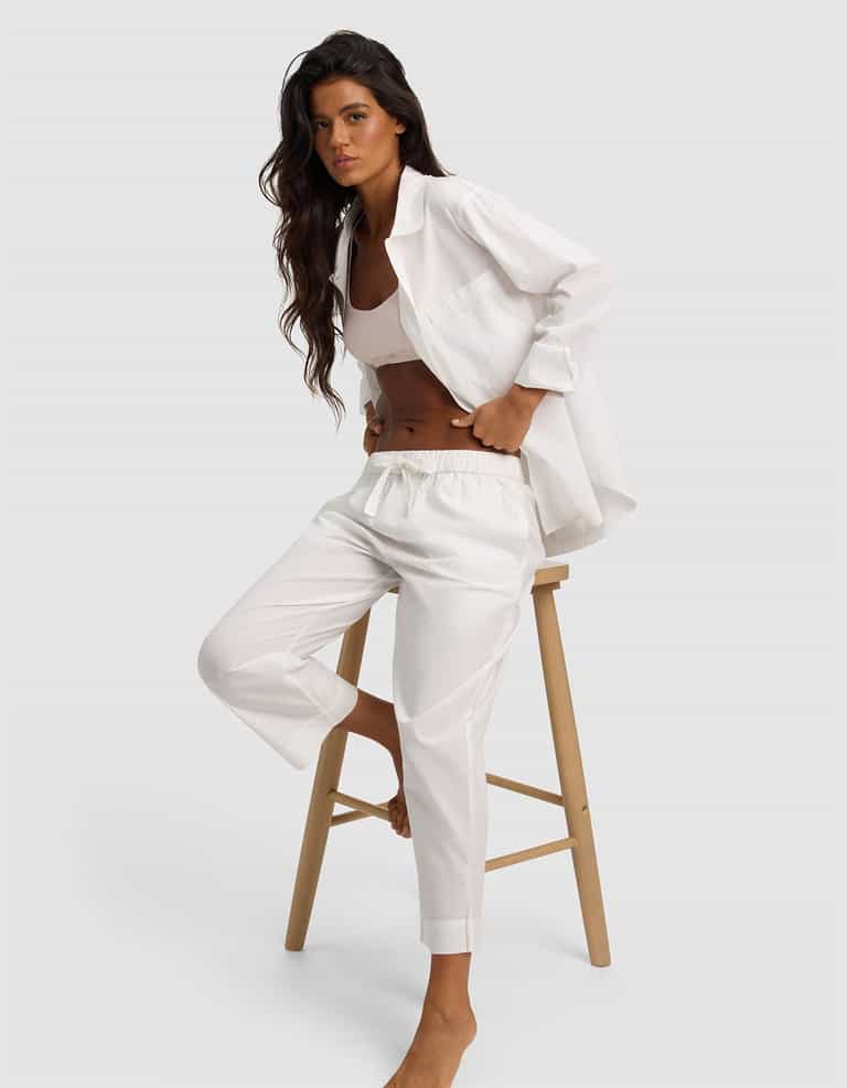 A woman with long dark hair sits on a wooden stool, wearing Cozy Earth’s Women's Sutton Cropped Pajama Pant in white with a matching loose shirt and crop top, posing relaxedly against a plain light gray background.|Color:White