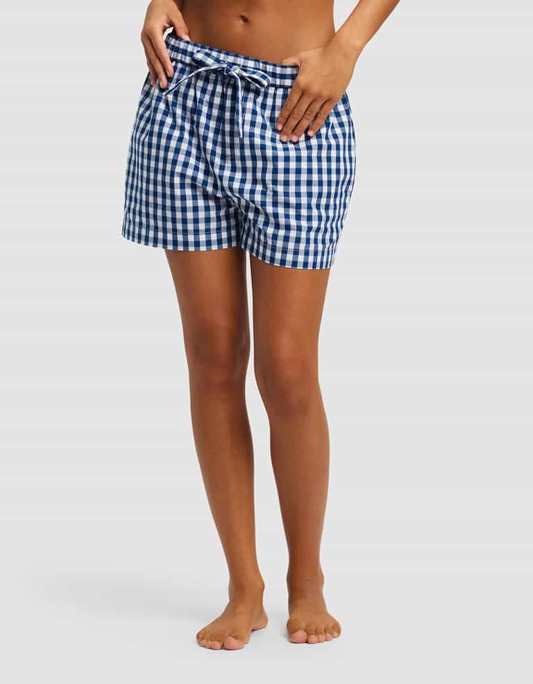 A woman stands barefoot against a plain light background, hands on hips, wearing Cozy Earth's Women's Sutton Pajama Short in blue and white gingham plaid with a drawstring waist.|Color:Pacific Gingham