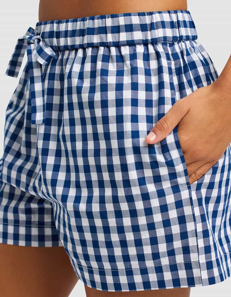 A close-up of someone wearing Cozy Earth's Women's Sutton Pajama Short in blue and white gingham, featuring an elastic waistband, a small bow, and one hand in the pocket.|Color:Pacific Gingham