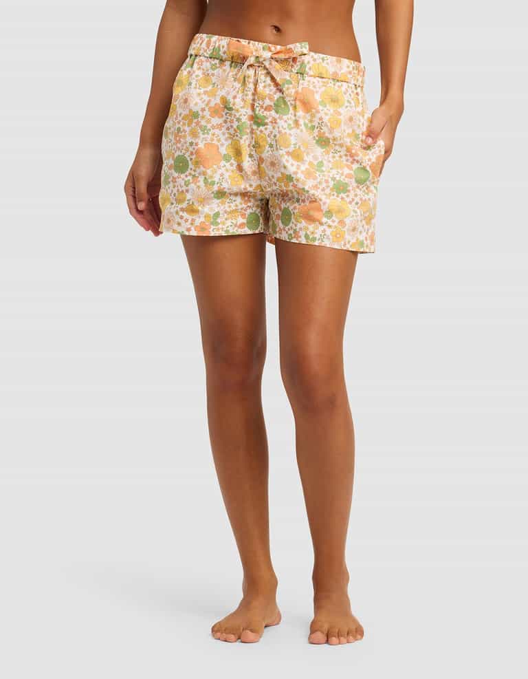 A person stands barefoot with hands in the pockets of Cozy Earth’s Women's Sutton Pajama Short, featuring a peach, green, and white floral print and a drawstring waist. Their torso and legs are shown against a plain light background.|Color:Wildflower